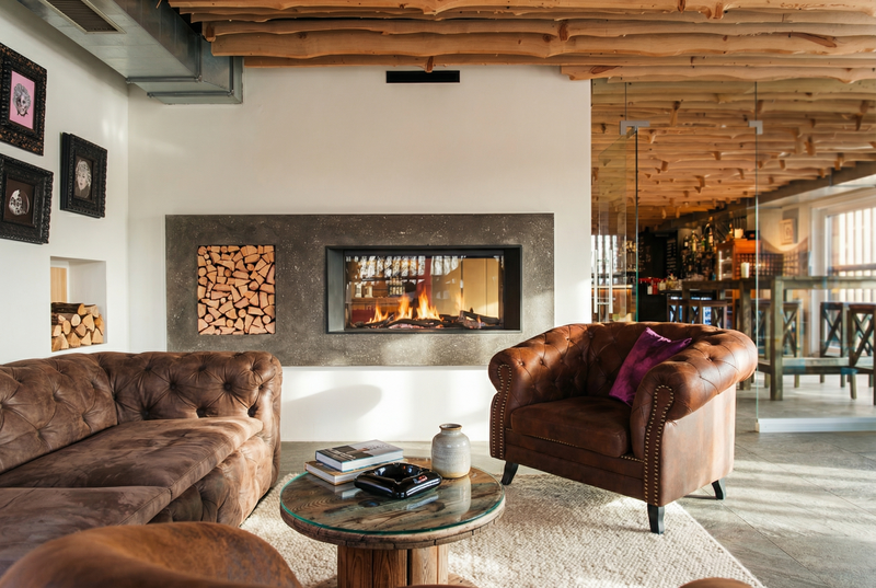 Diese Feuerstelle integriert moderne Technik mit rustikalen Elementen. Die Betonverkleidung rahmt den Kamineinsatz und die Holzlagerung harmonisch ein. Die lebendigen Flammen und das gestapelte Holz sorgen für eine warme Atmosphäre.